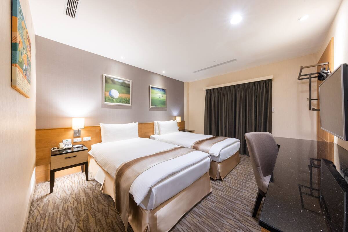 A hotel room with two beds, a desk, a chair, a TV, and artwork on the walls.