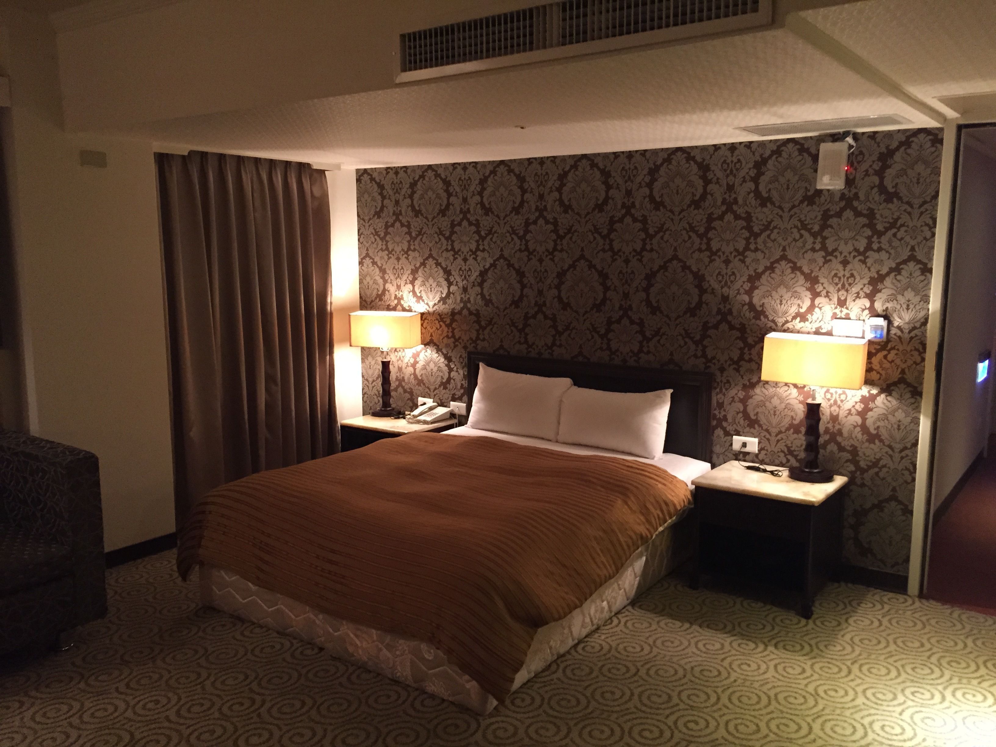 Una habitación de hotel con una cama grande, dos mesitas de noche con lámparas, un teléfono y un papel tapiz estampado.