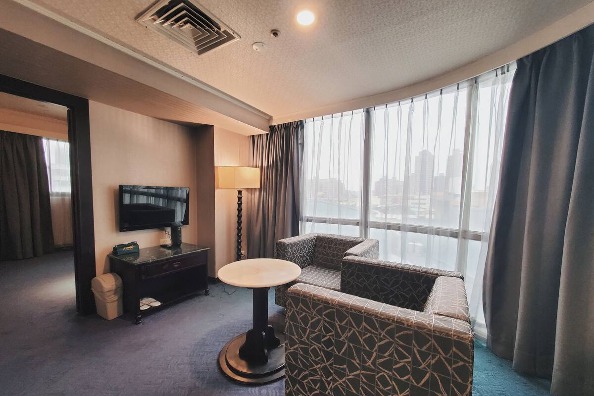 Une chambre d’hôtel dotée d’une grande fenêtre, d’un téléviseur à écran plat, d’une petite table ronde, de fauteuils à motifs et d’une commode de couleur sombre.