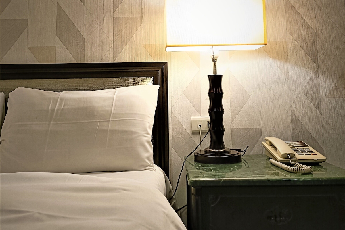 Una cama bien tendida con sábanas blancas, una lámpara de noche, un teléfono y una mesita de noche verde.