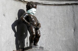 Une statue en bronze représentant un enfant portant un masque sur la tête, et qui se tient sur un piédestal devant un mur en béton.