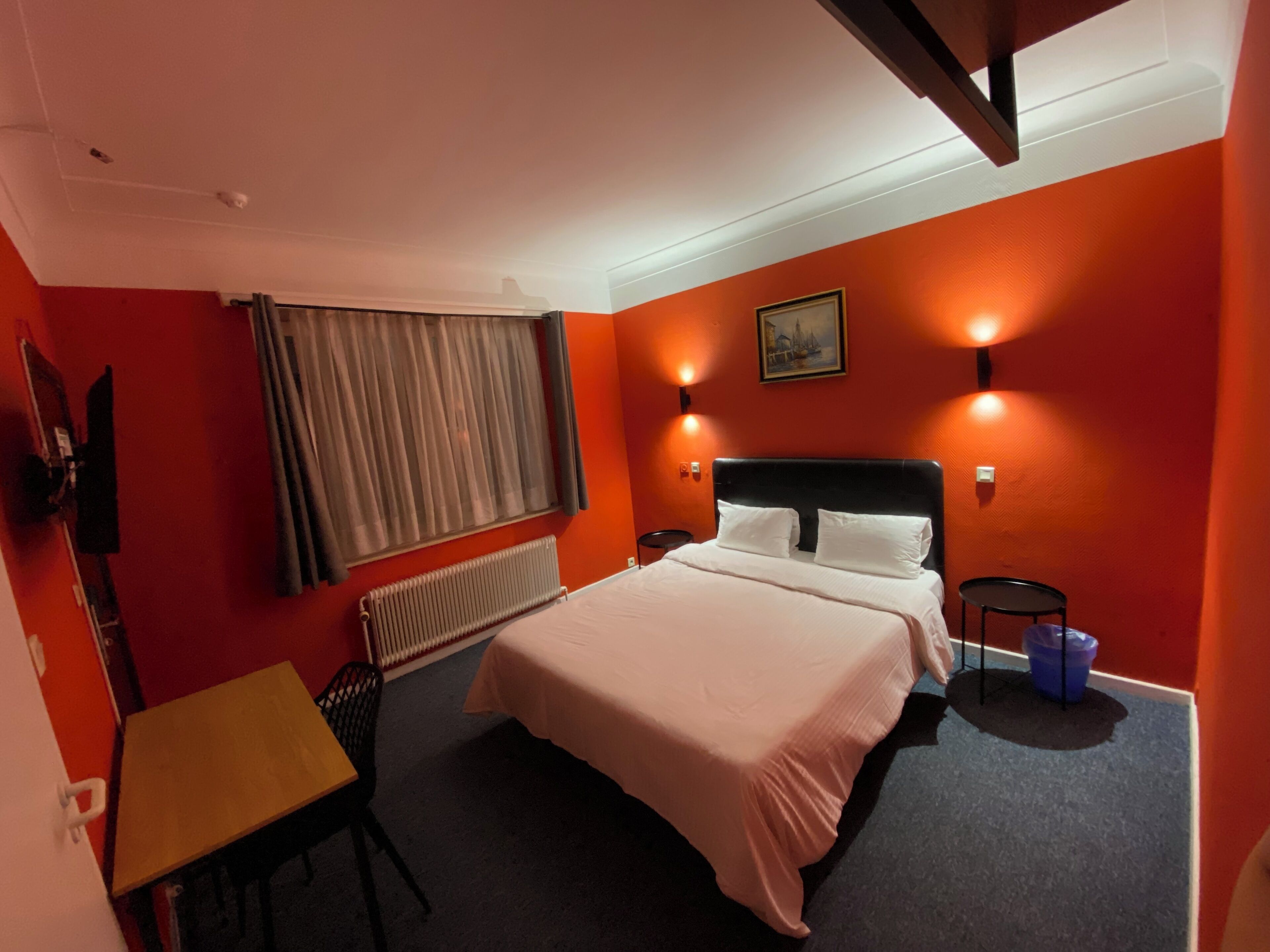 Quarto de hotel com cama, poltrona, mesa, radiador e um quadro na parede.