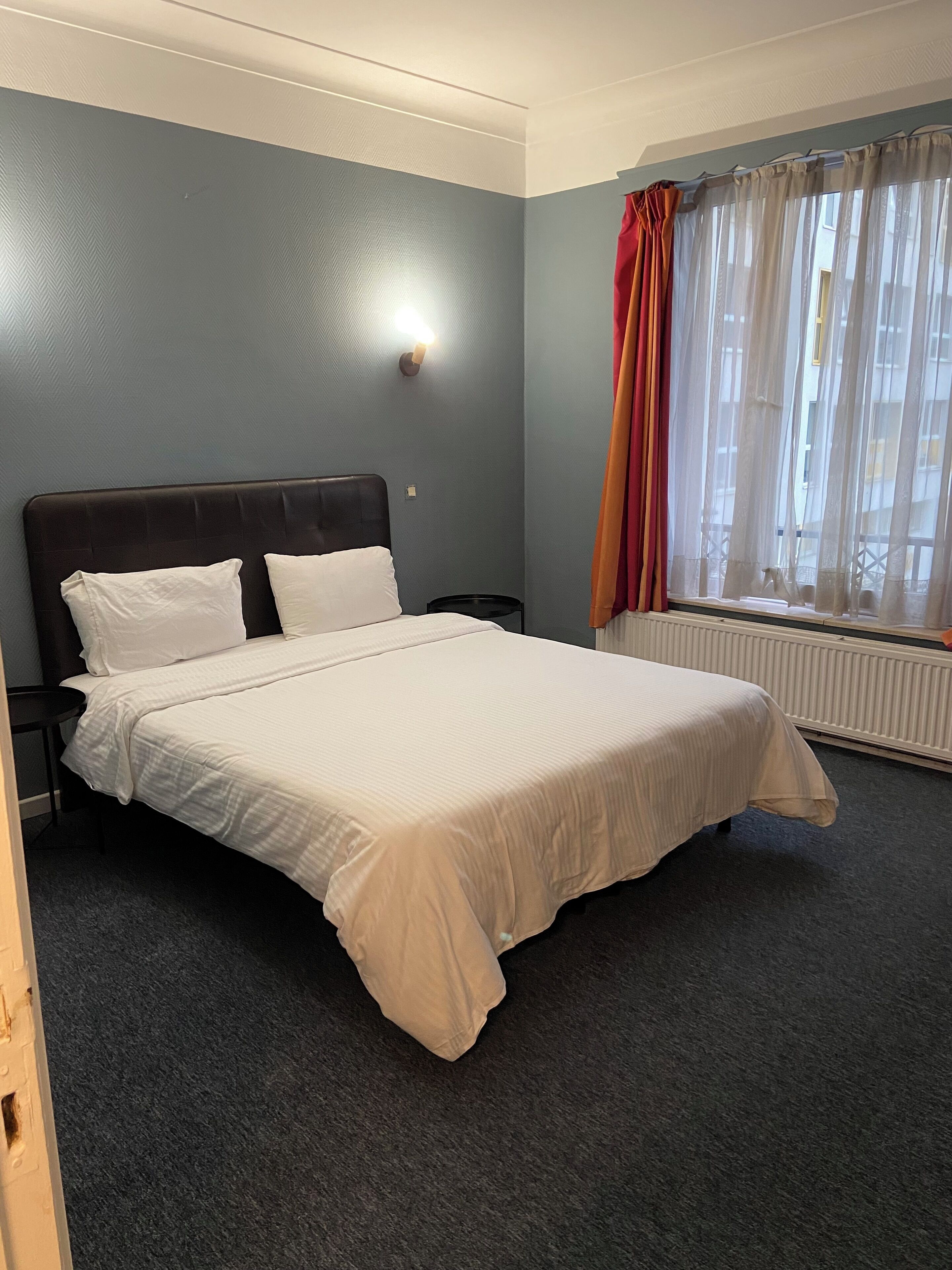 Een hotelkamer met een bed, nachtkastje, wandlamp, raam met gordijnen en een radiator.
