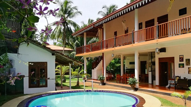 Un edificio de dos plantas con piscina, mobiliario exterior y abundante vegetación.