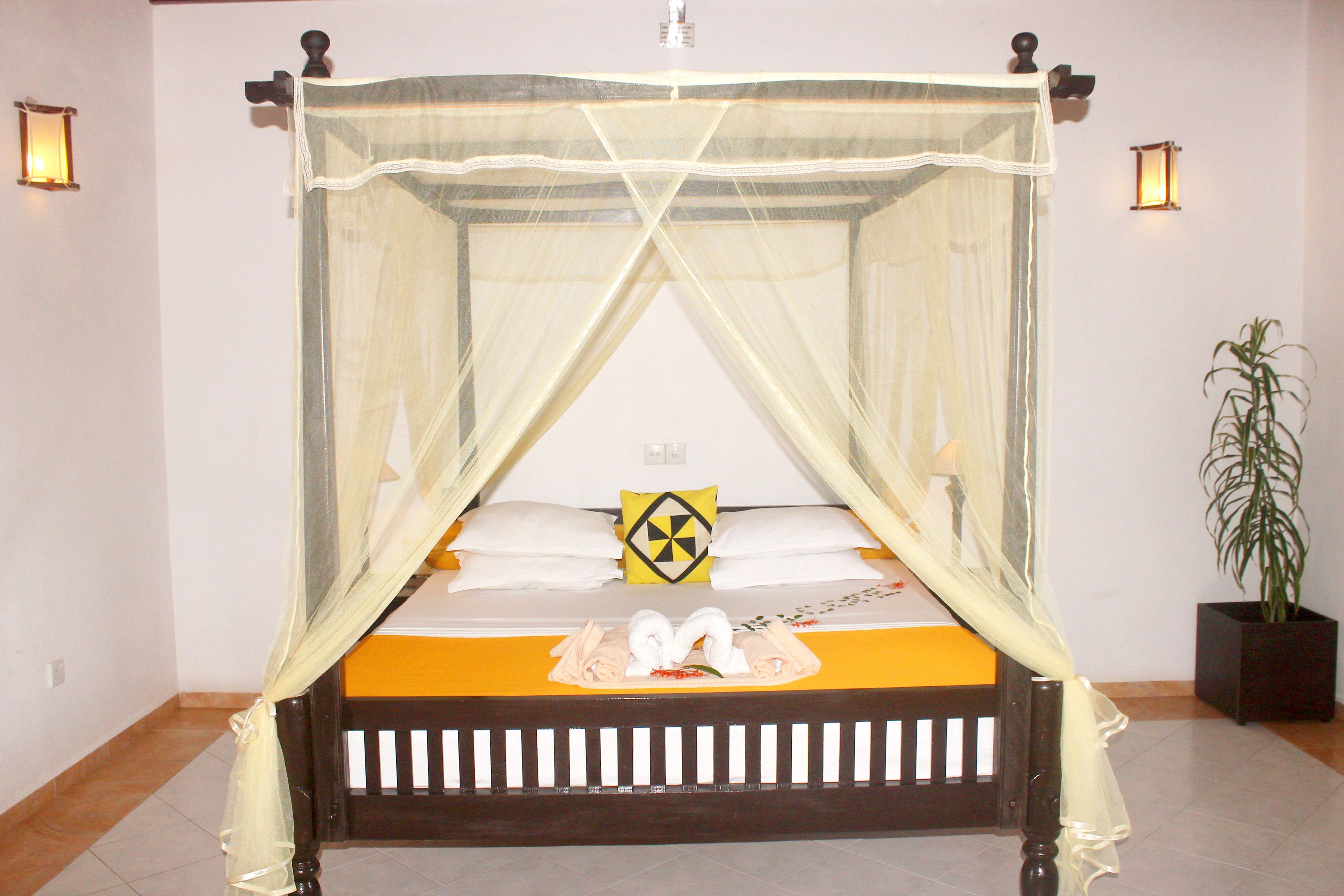 Una cama con dosel y cuatro columnas, con un edredón amarillo y almohadas blancas, una almohada decorativa con estampado negro y amarillo, y una planta en maceta a la derecha.
