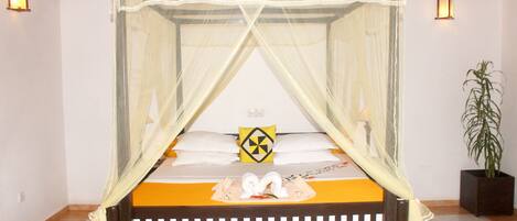 Una cama con dosel y cuatro columnas, con un edredón amarillo y almohadas blancas, una almohada decorativa con estampado negro y amarillo, y una planta en maceta a la derecha.