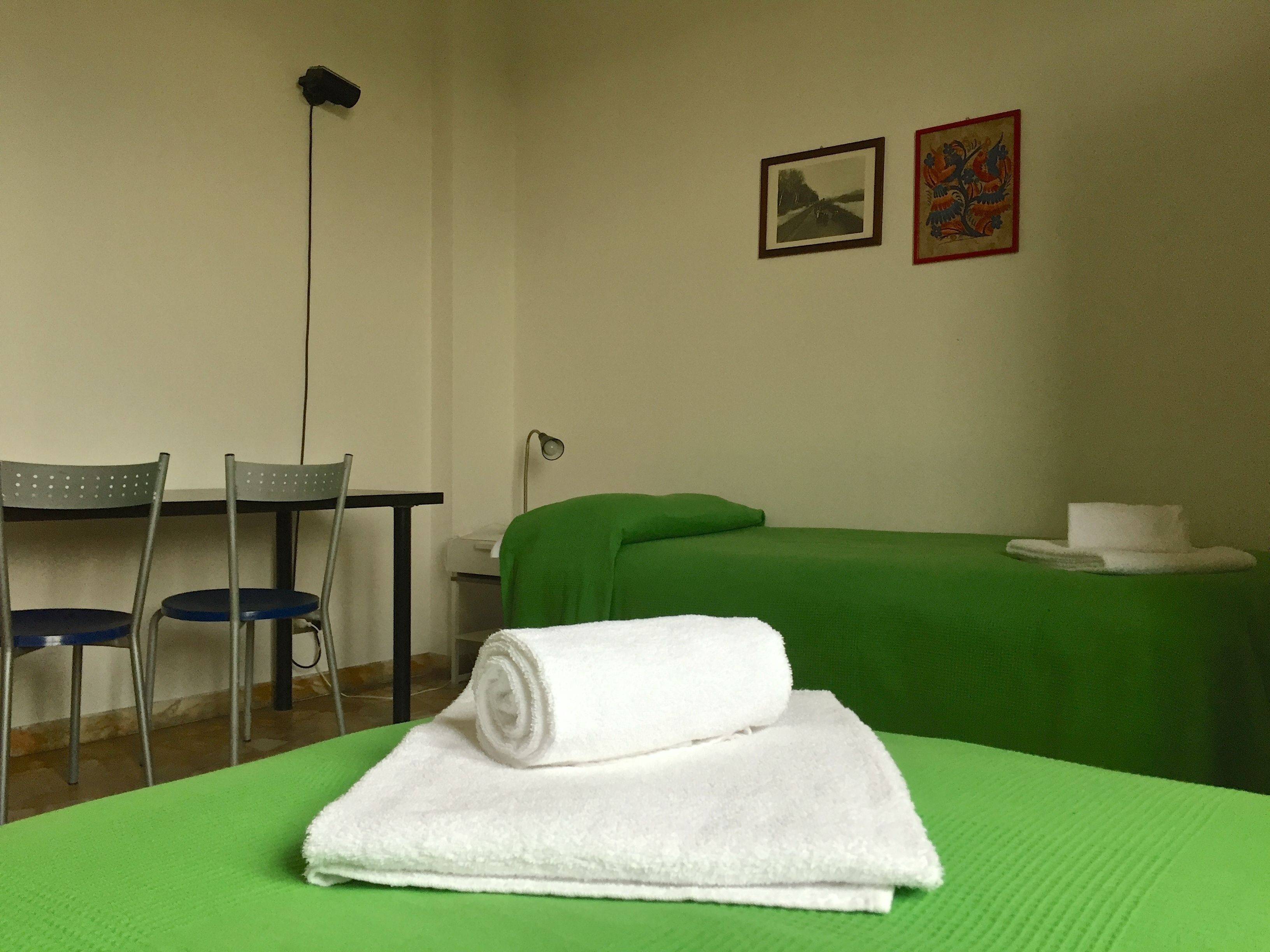 A room with a green bed, a white towel, two chairs, a desk, and two framed pictures on the wall.