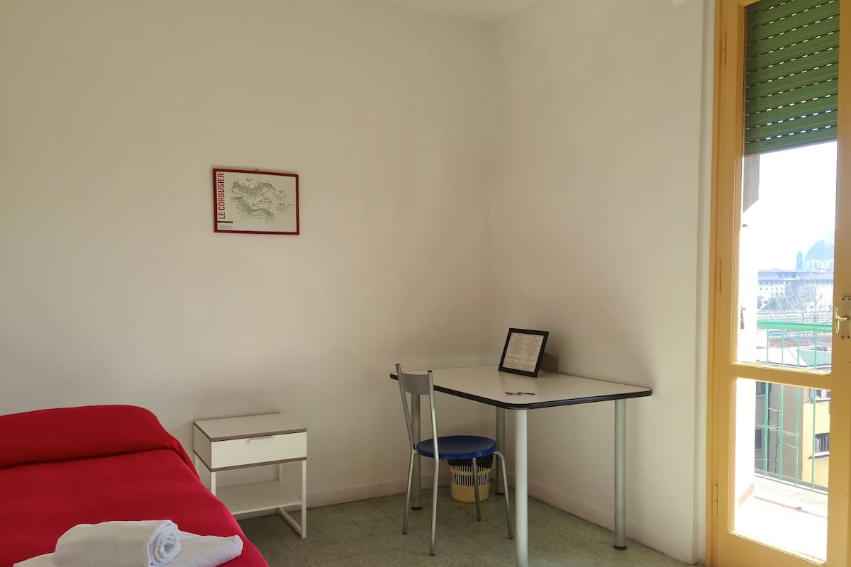 A small, bright room with a bed, a desk, and a chair. There is a window with a view of buildings outside.