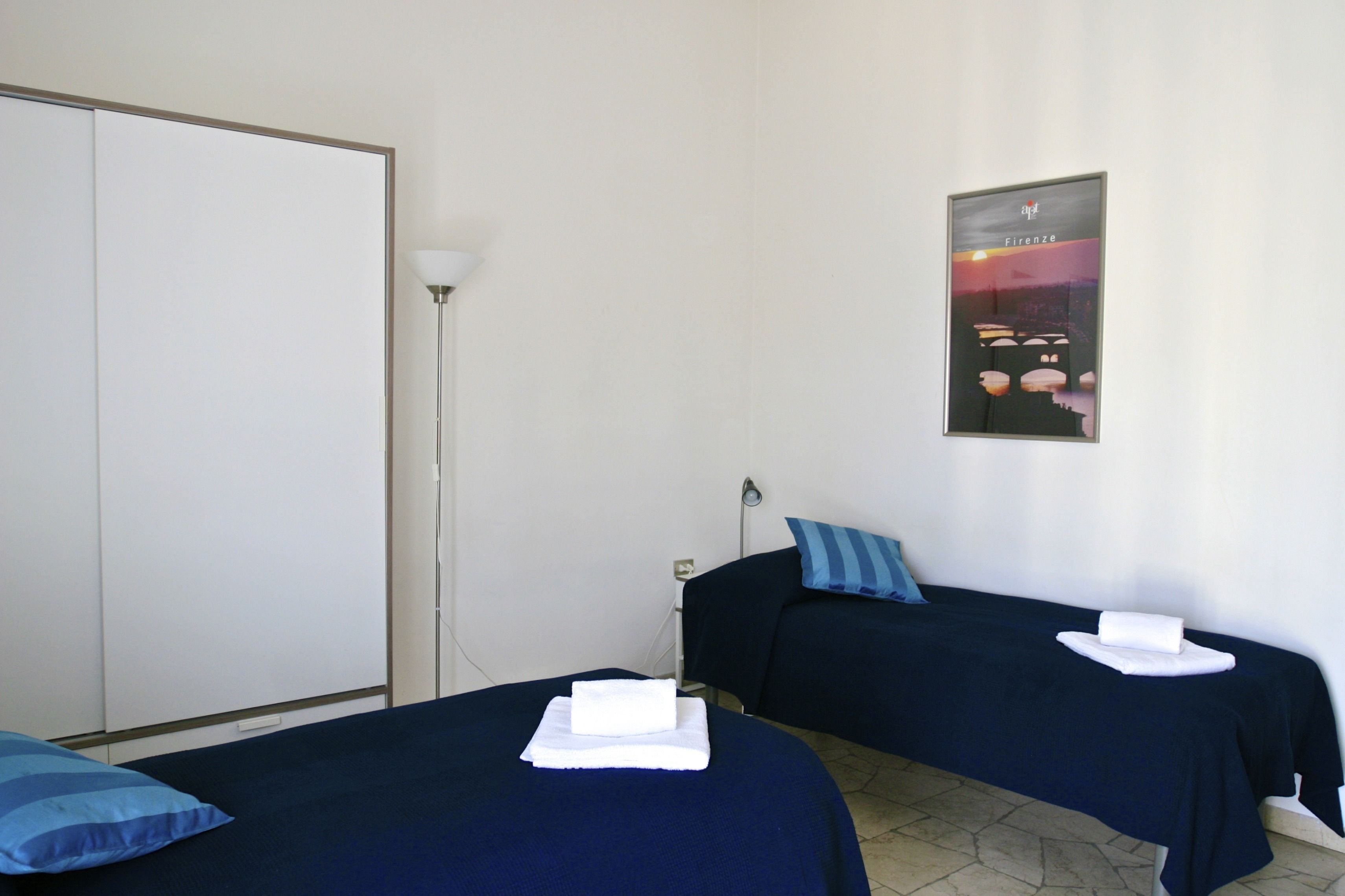 A hotel room with two beds, each with a blue bedspread and a white towel. There is a poster on the wall and a floor lamp.