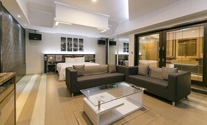 A modern hotel room with a large bed, two sofas, a glass coffee table, and a sliding glass door leading to a balcony.