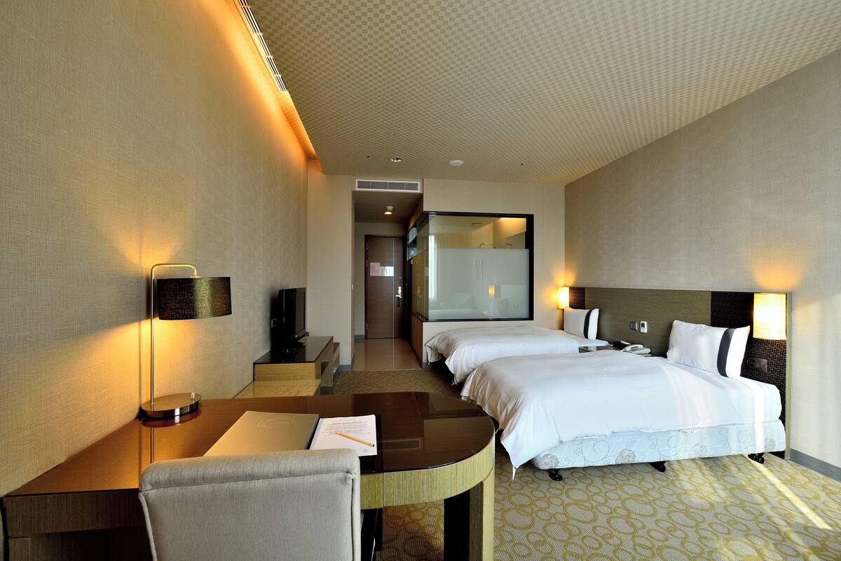 A hotel room with a desk, two beds, a chair, and a large mirror.