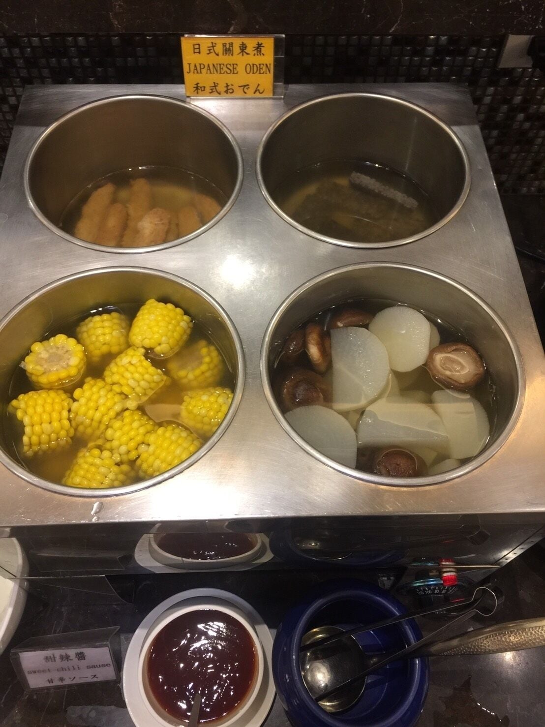 A kitchen with four pots containing different foods, a sign indicating Japanese Oden, and a bowl of sweet chili sauce.