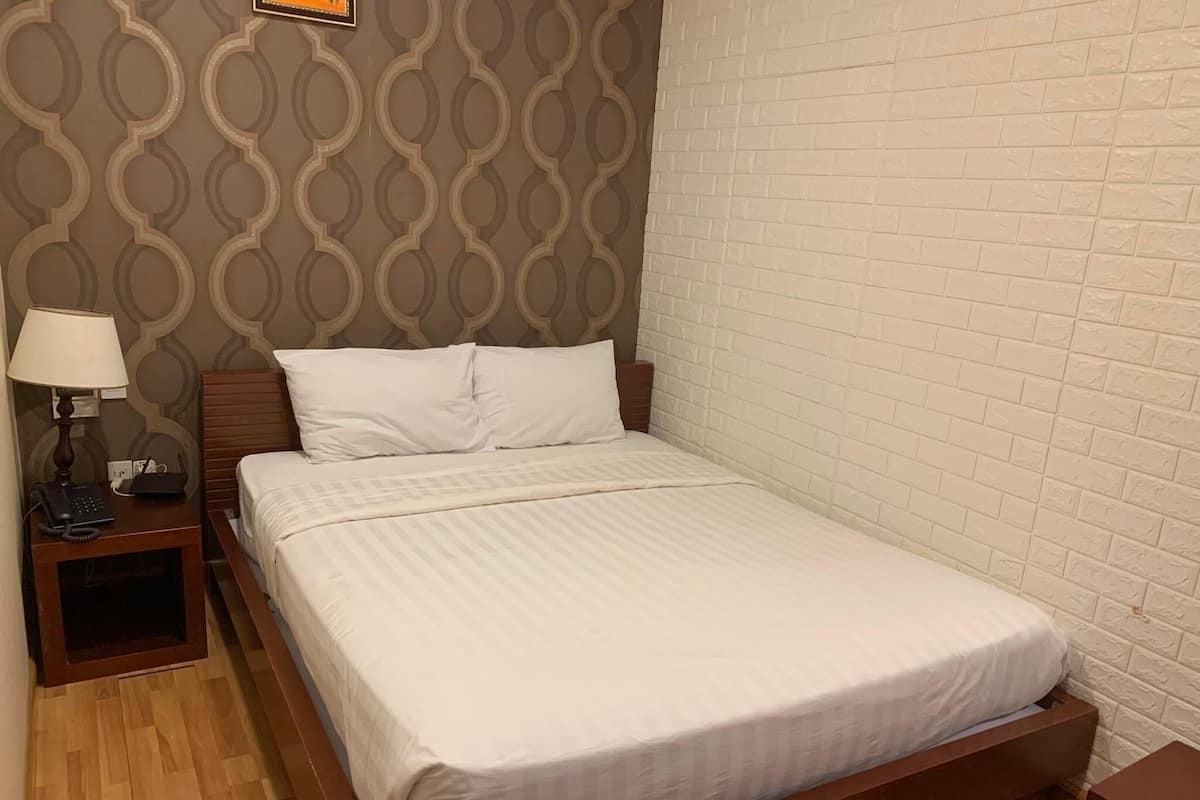 A neatly made bed in a hotel room with a patterned wallpaper, a wooden nightstand with a lamp, and a brick-patterned wall.