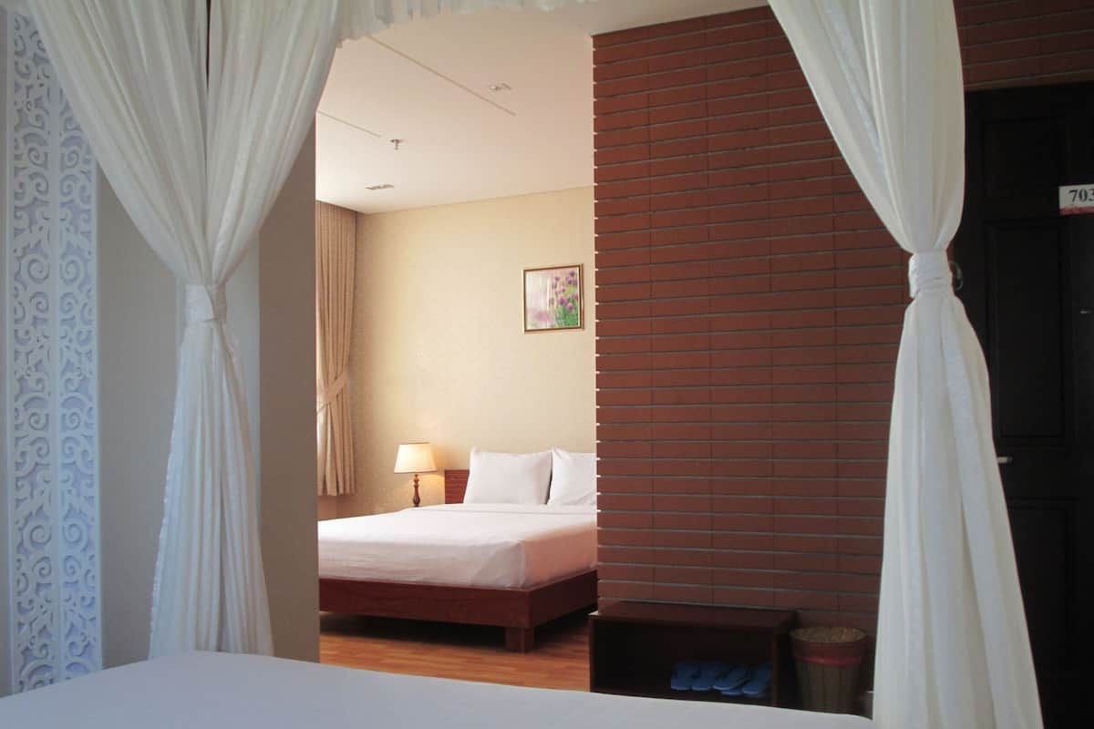 A hotel room with a bed, a nightstand with a lamp, a small table, and a door with a number sign.