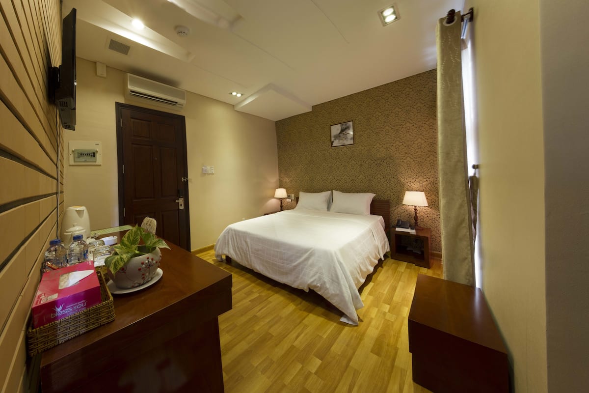 A hotel room with a bed, bedside lamps, a wooden floor, a desk with a basket of toiletries, and a wall-mounted TV.