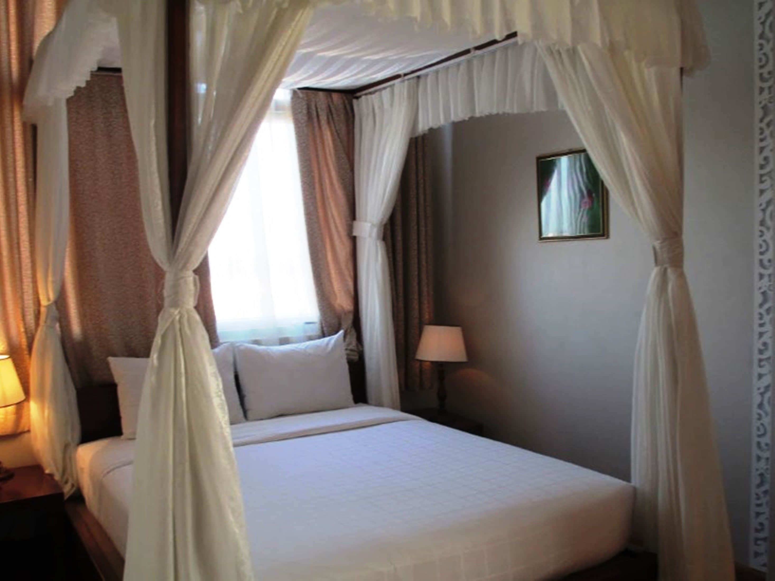 Una habitación de hotel con cama, cortinas, mesita de noche, lámpara y un cuadro en la pared.