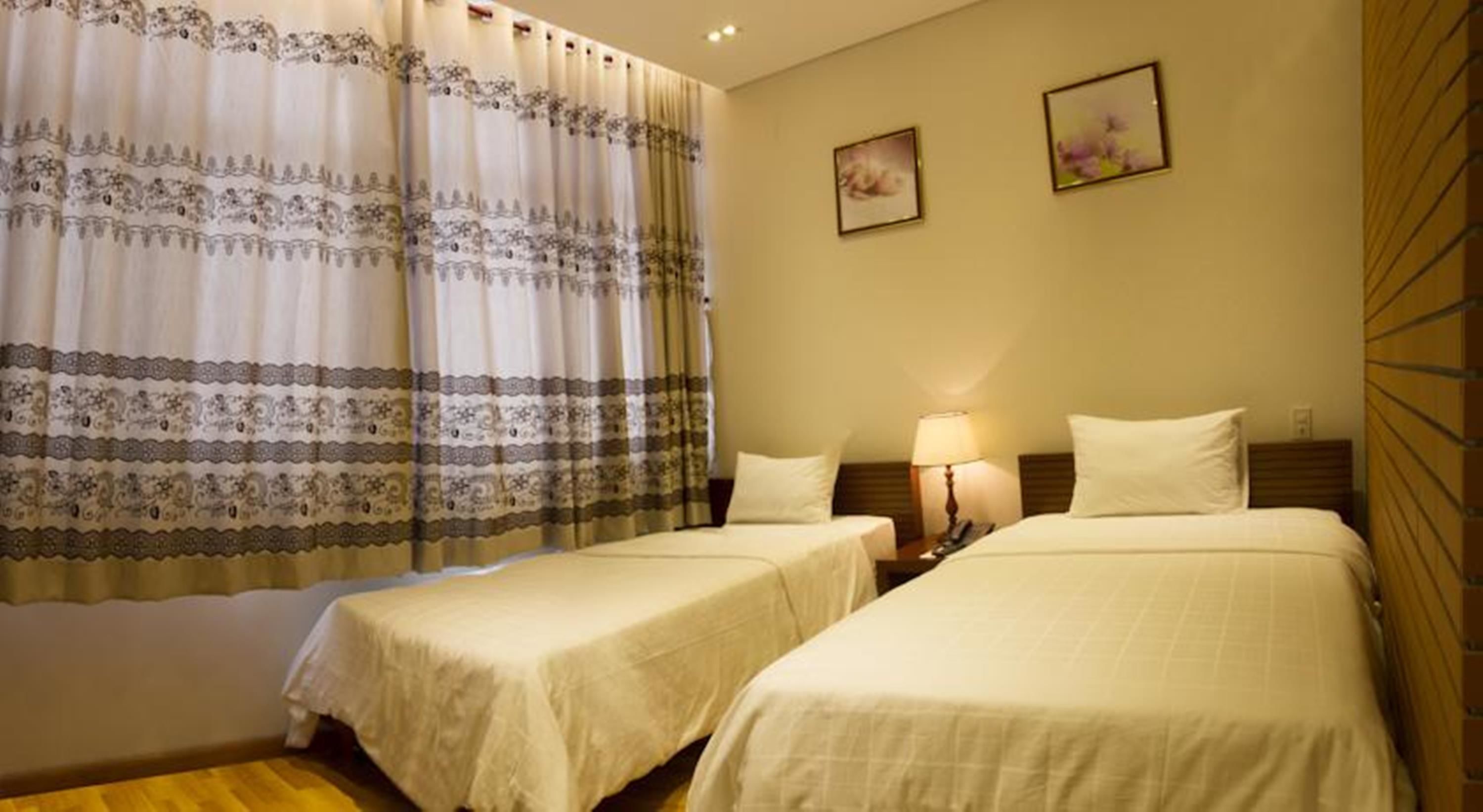 Habitación de hotel con dos camas individuales, una mesita de noche con lámpara y un ventanal con cortinas estampadas.