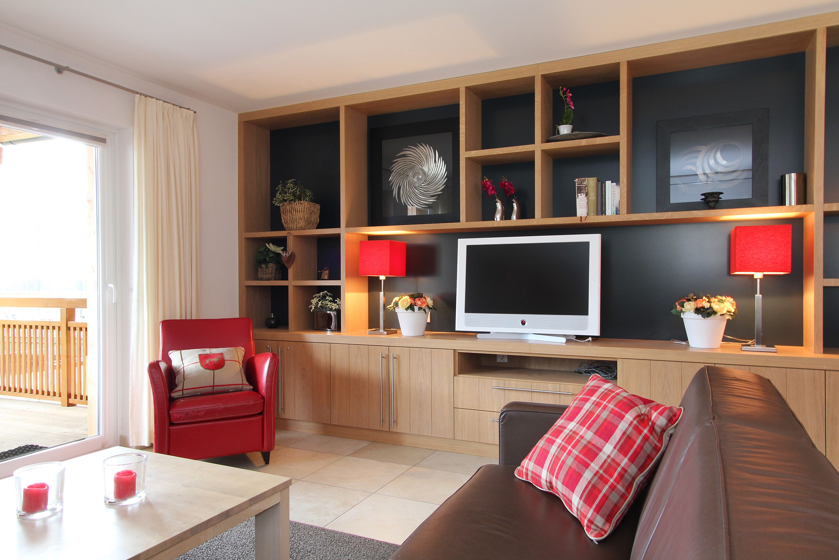 A modern living room with a flat-screen TV, red and white decor, and a wooden shelving unit.