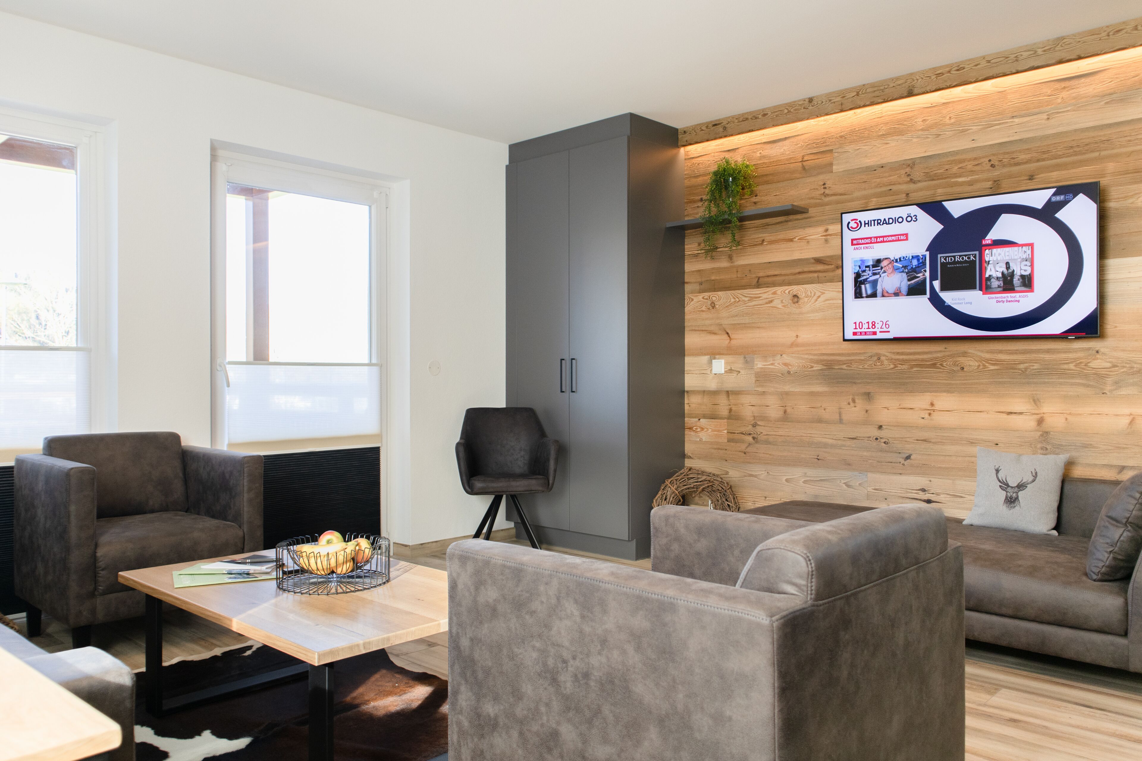 Una sala de estar moderna con sofás grises, un muro de acento de madera y un televisor montado que muestra un programa.