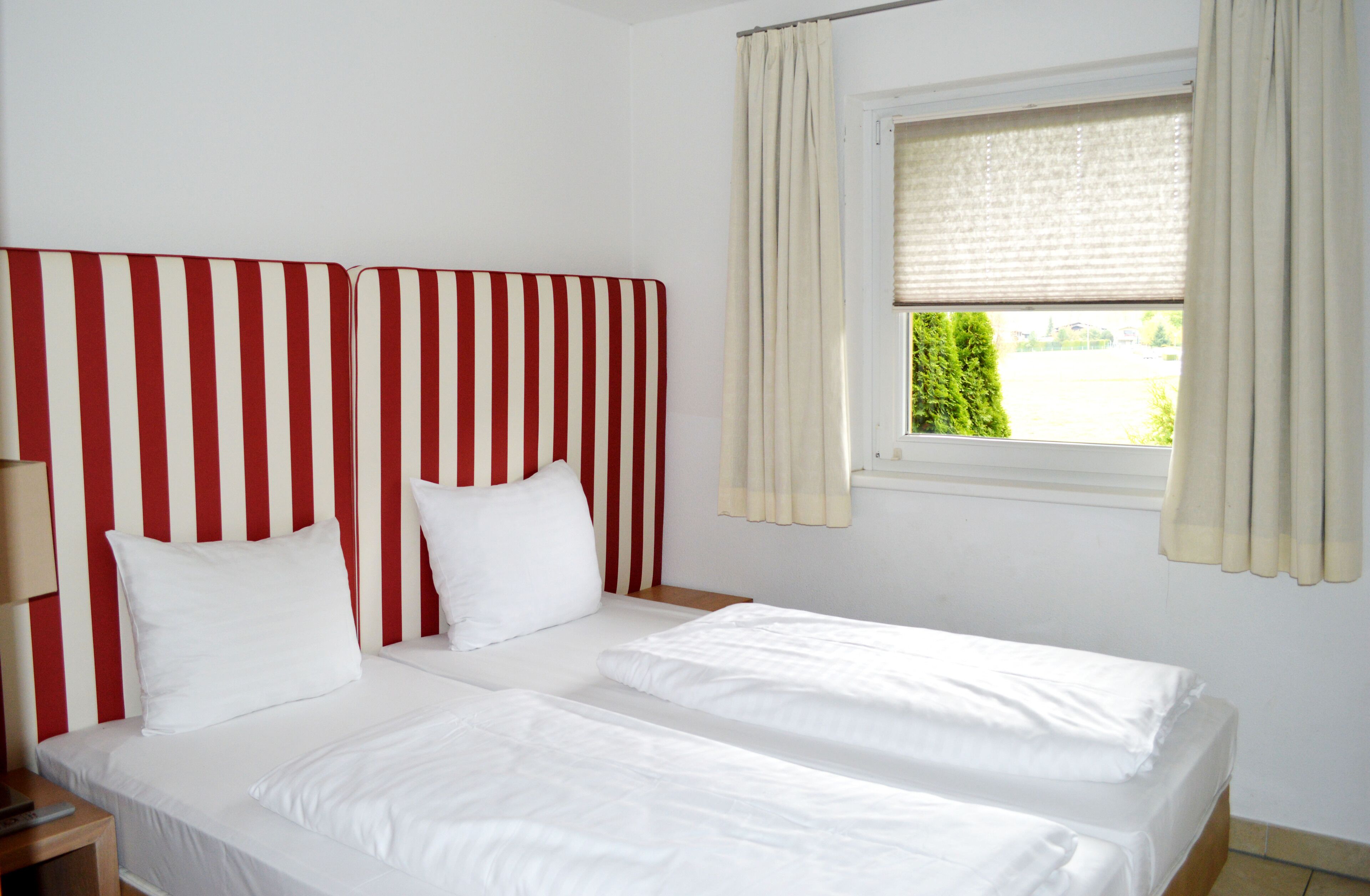 Une chambre d’hôtel avec une tête de lit à rayures, deux lits, une fenêtre avec des rideaux et une table de chevet avec une lampe.