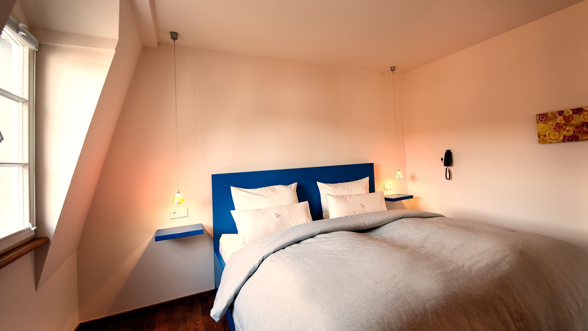 Ein Schlafzimmer mit schrägem Dach, einem Bett mit blauer Kopfteil, weißer Bettwäsche, einem Nachttisch mit Lampe und einem an der Wand montierten Telefon.