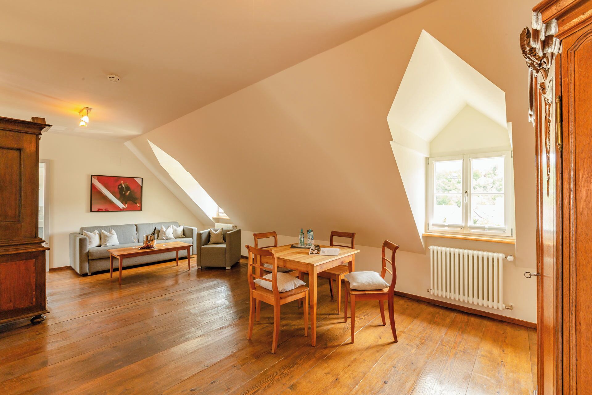 A room with a sloped ceiling, wooden flooring, a dining table with chairs, a sofa, and a radiator.