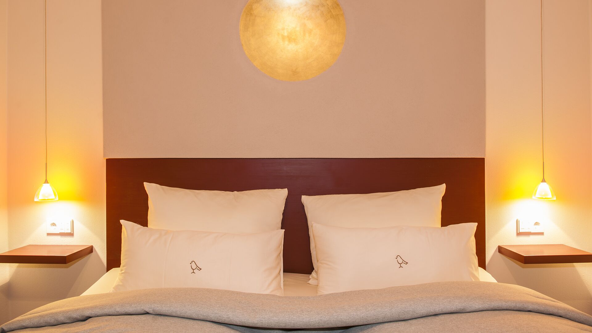 Quarto de hotel com uma cama de casal, duas mesas de cabeceira, luminária fixada na parede e um pequeno estante em cada mesa de apoio.