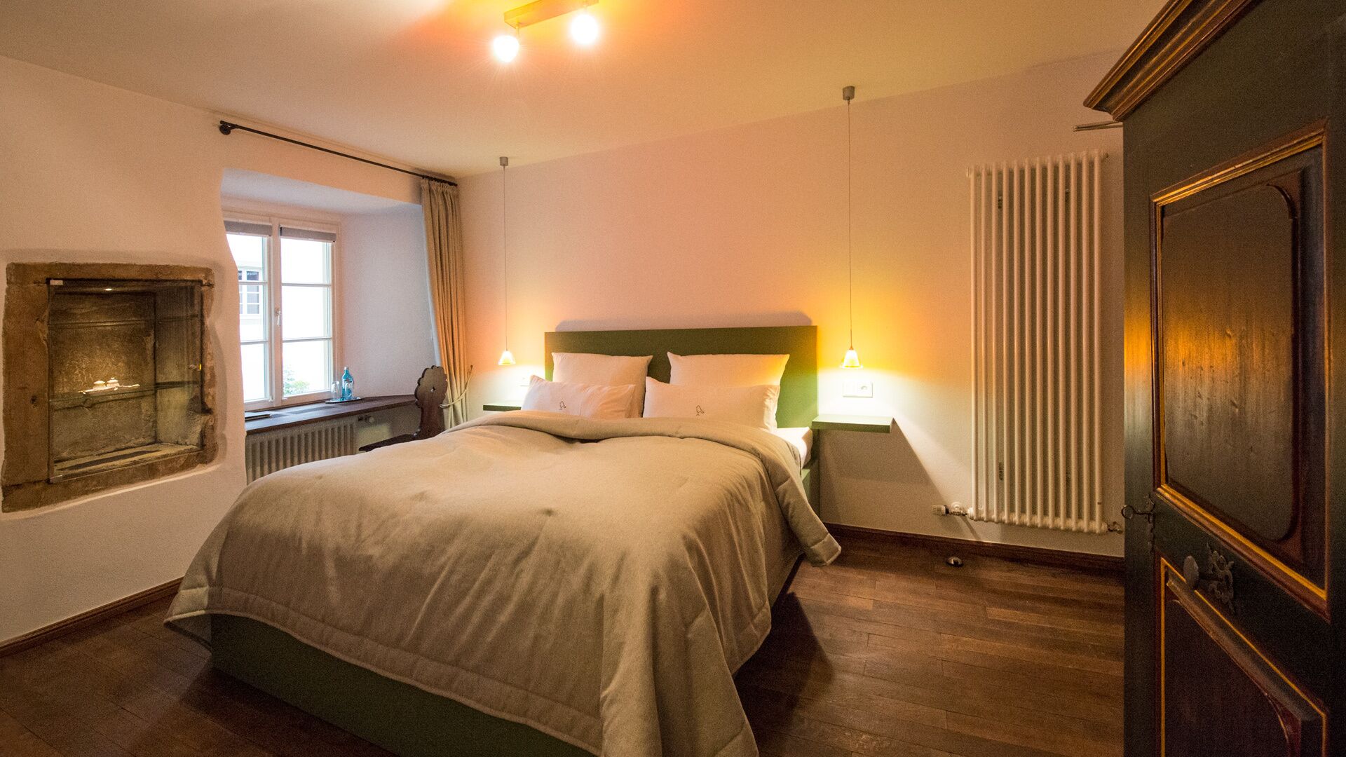 A hotel room with a large bed, bedside lamps, a radiator, and a window with curtains.