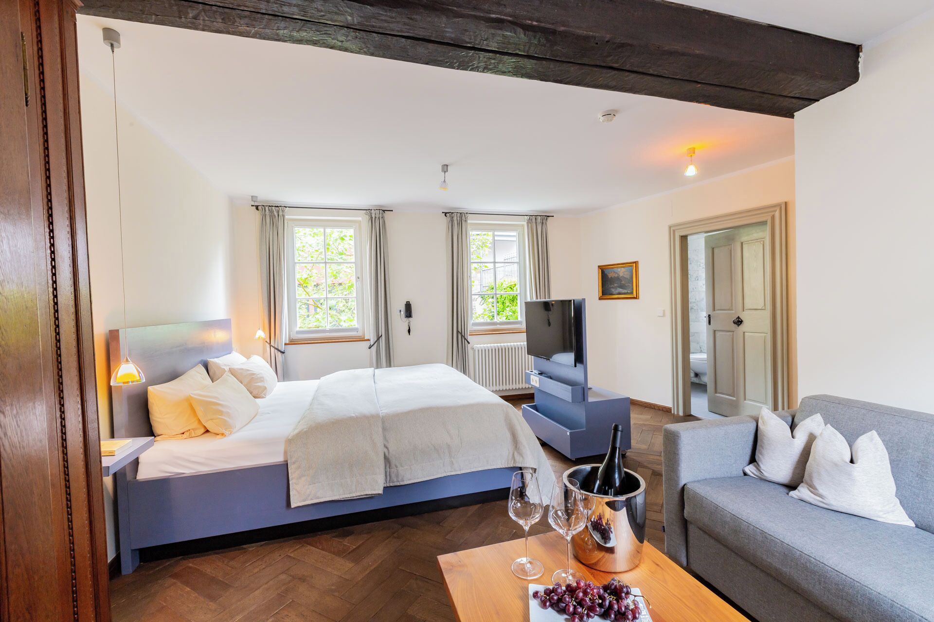 A hotel room with a bed, a television, a sofa, and a table with wine glasses and grapes.