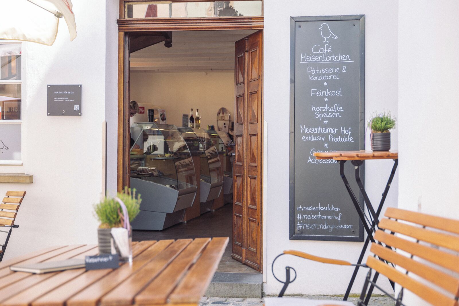 An outdoor café with wooden tables and benches, a chalkboard menu, and a glass display case with pastries.