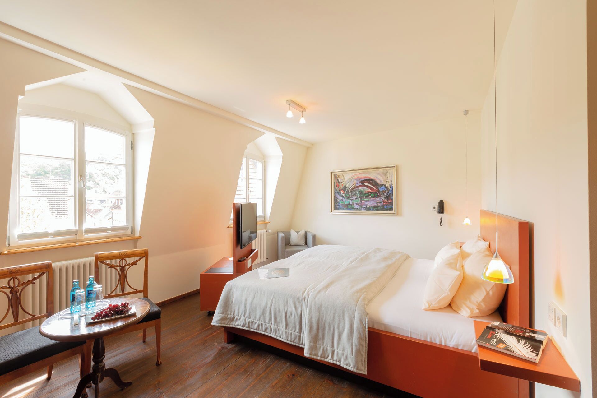 Habitación de hotel con cama, escritorio, silla, mesa con botellas y uvas, y ventana con vista a edificios.