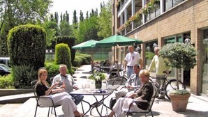 Un patio al aire libre con mesas y sillas, rodeado de vegetación y un edificio con ventanas decoradas con flores.