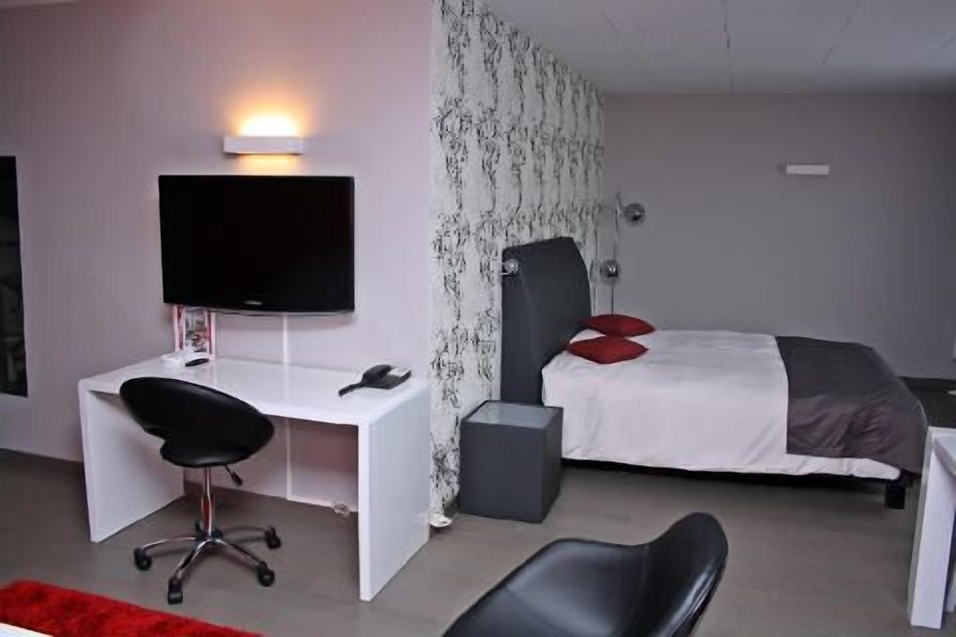 A modern bedroom with a white desk, black office chair, a bed with red pillows, and a TV mounted on the wall.