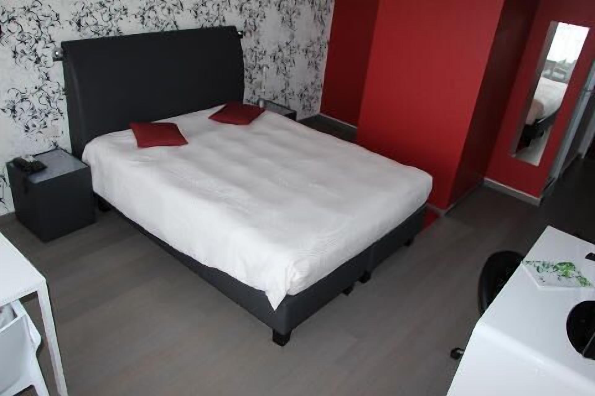 A modern bedroom with a large bed, a nightstand, a desk, and a red accent wall.