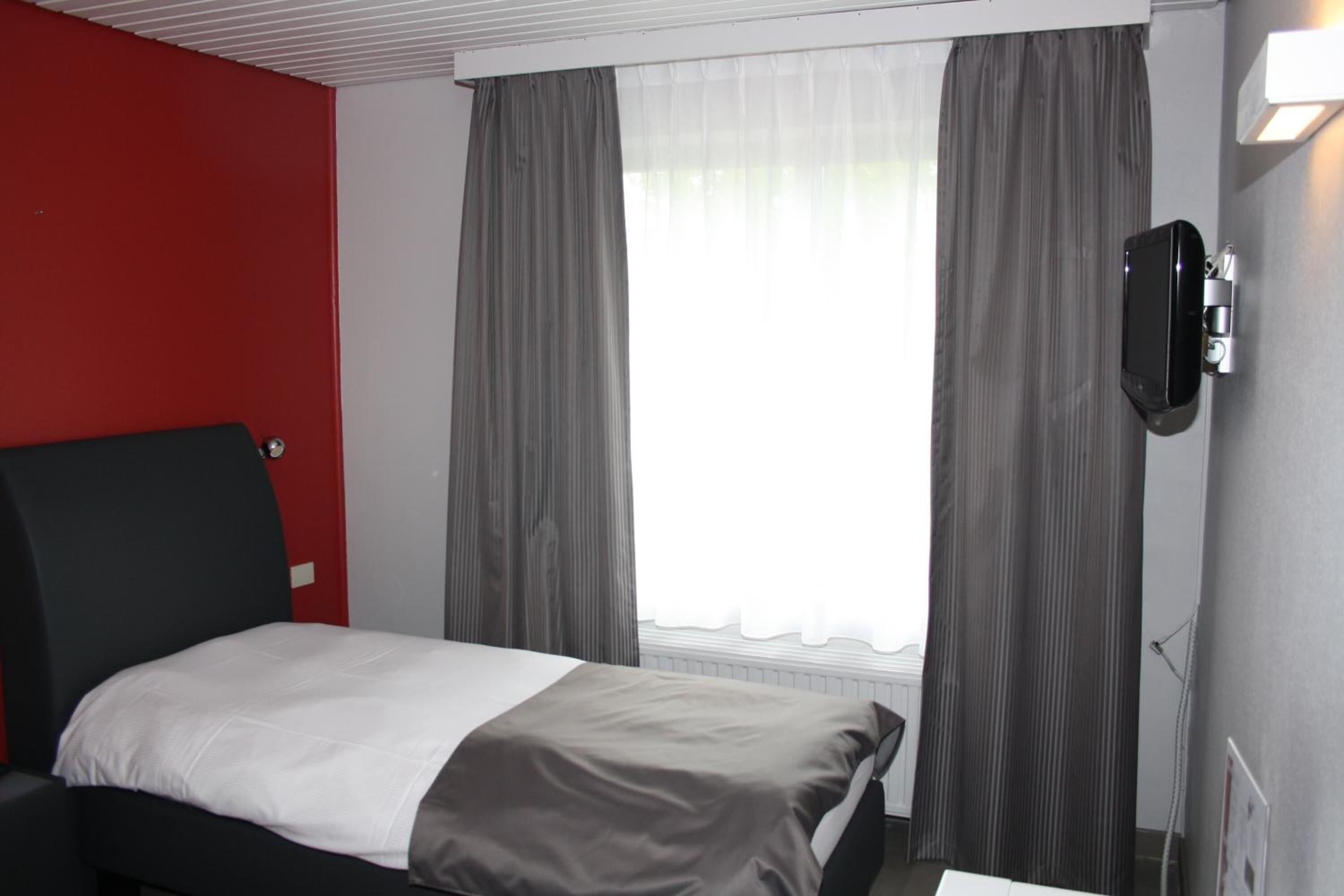 Une chambre d’hôtel avec un lit, un mur d’accent rouge, des rideaux gris et un téléviseur fixé au mur.