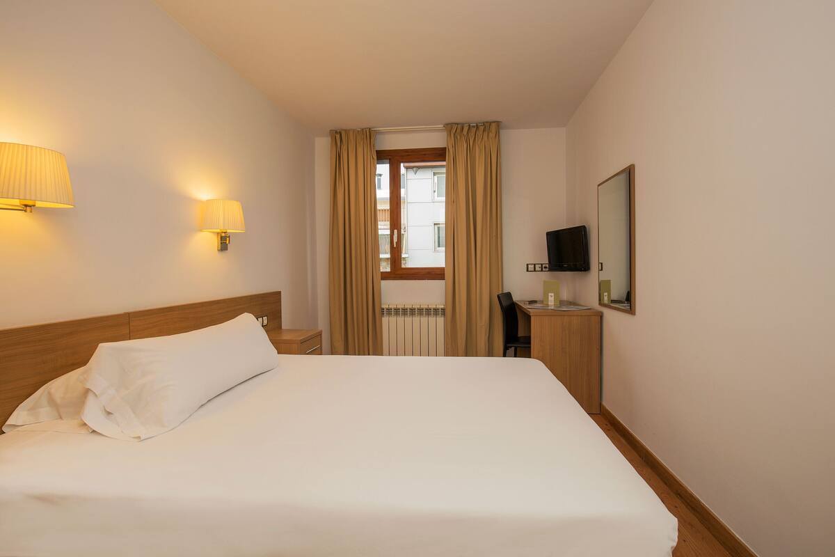 Habitación de hotel con cama, escritorio, silla, televisor y ventana con cortinas.