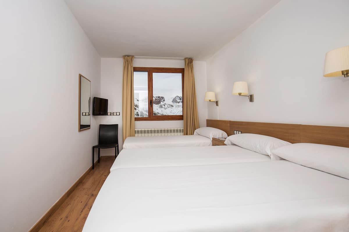 Habitación de hotel con piso de madera, cama con sábanas blancas, una silla, televisor y ventana con cortinas.
