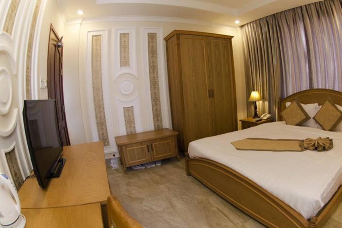 A hotel room with a large bed, wooden wardrobe, bedside tables with lamps, a desk with a chair, and a television.
