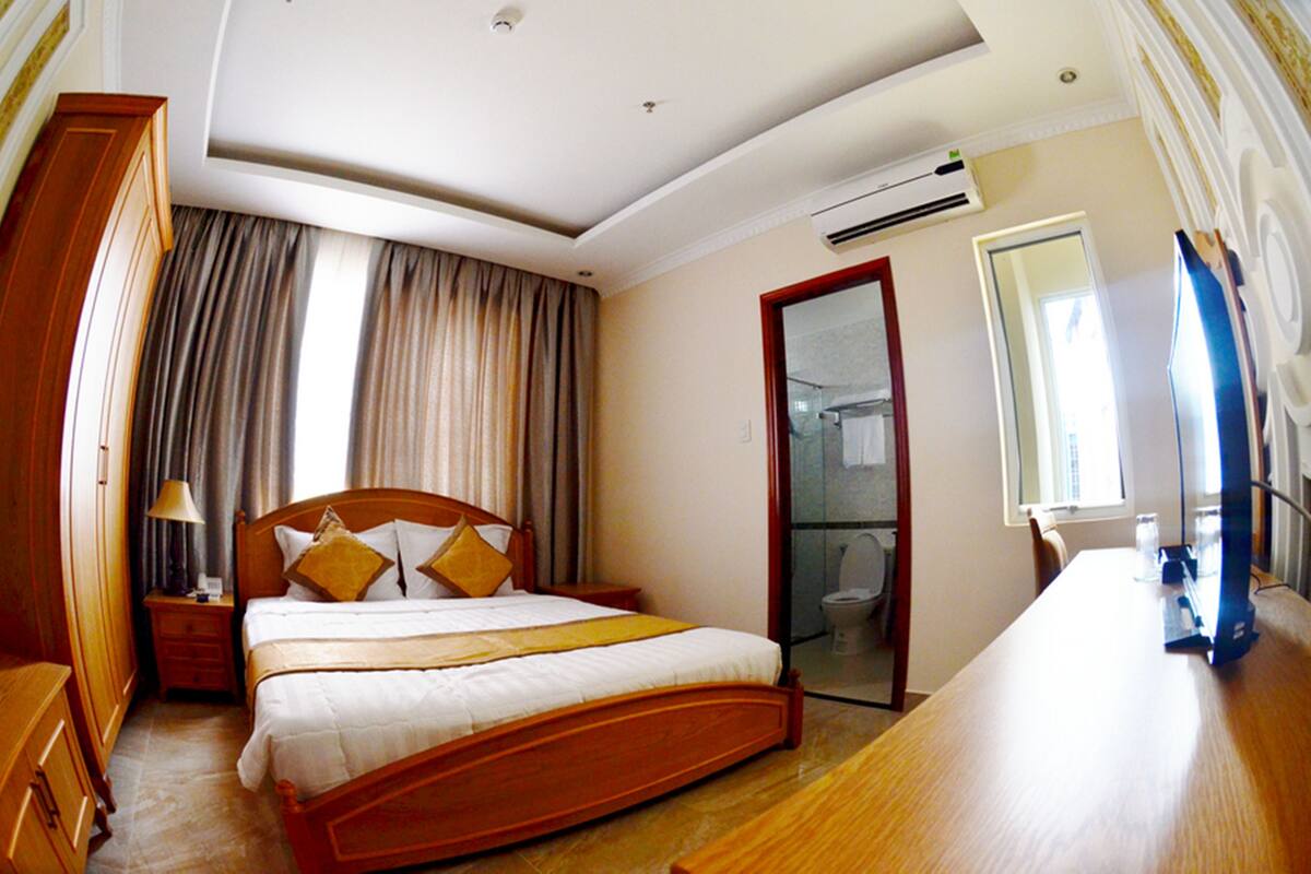 A hotel room with a wooden bed, a desk, a mirror, and a bathroom visible through a glass door.