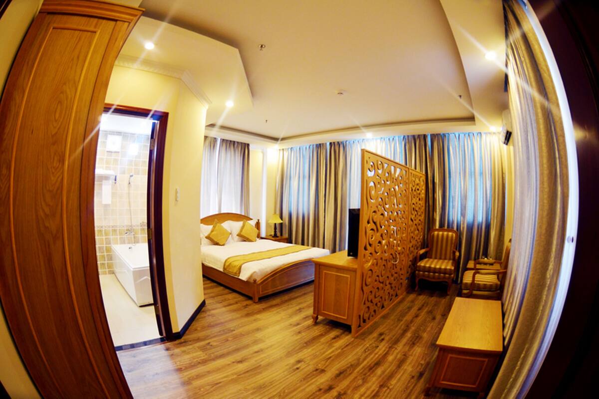 A hotel room with a wooden floor, a bed with a headboard, a desk, a chair, and a partition with a decorative pattern.