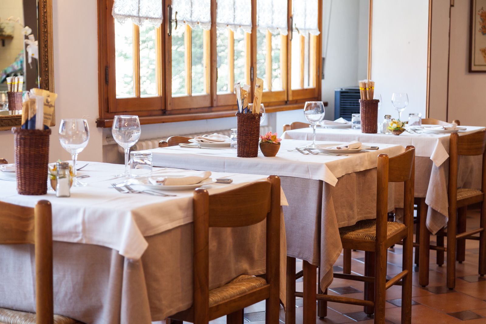 A dining area with tables set for two, wooden chairs, and a window with lace curtains.