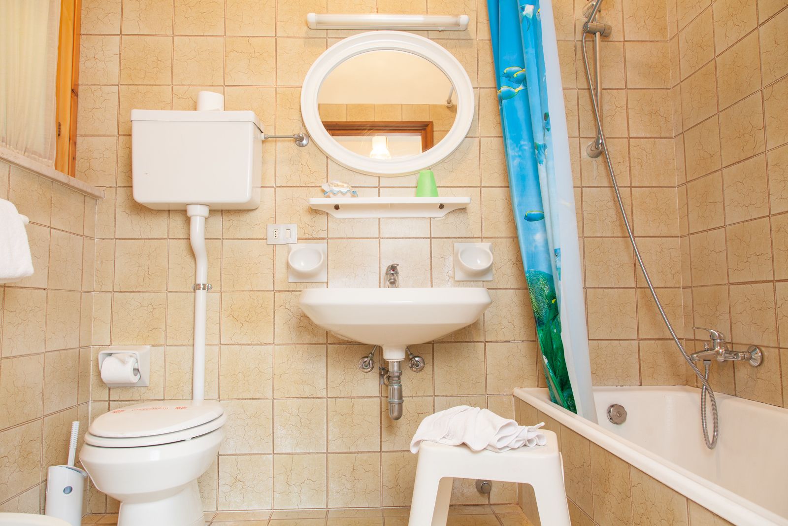 A bathroom with a toilet, sink, mirror, shower, and a small stool.
