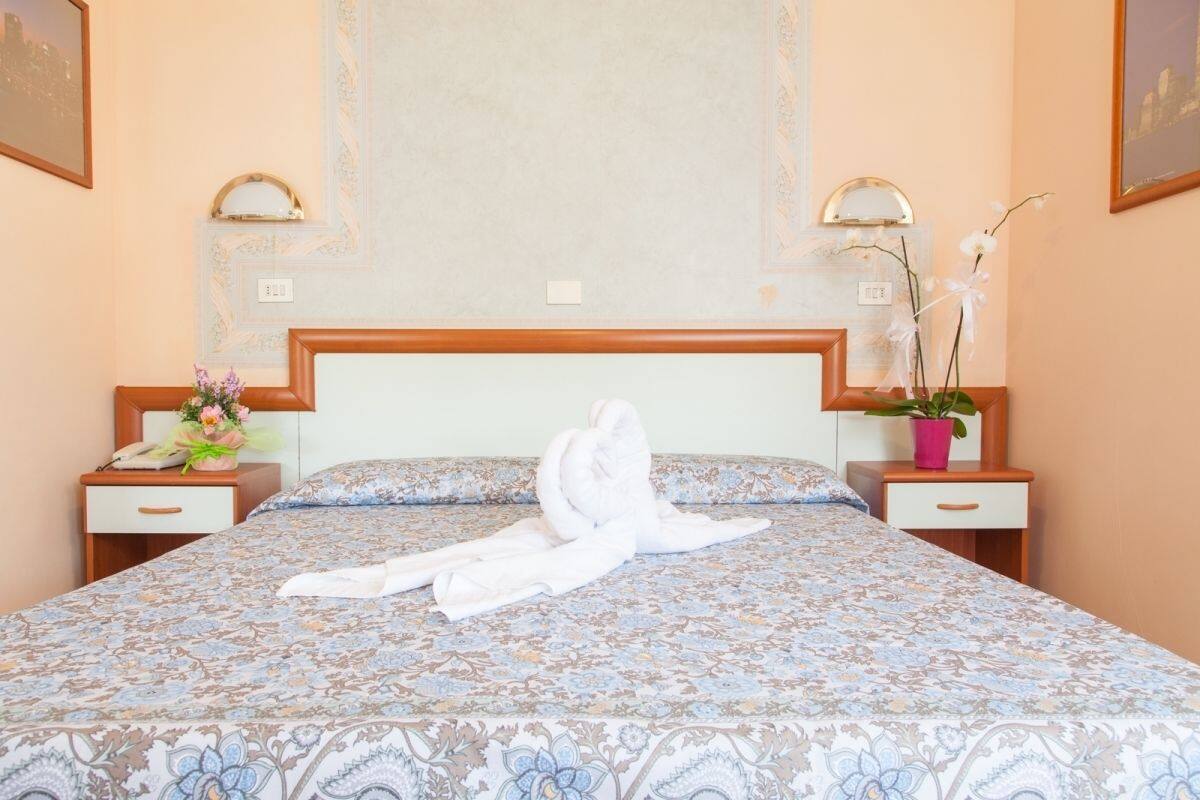 Une chambre d’hôtel avec une couvre-lit à motifs floraux, une tête de lit en bois, deux tables de chevet avec des lampes, une petite table de nuit avec un téléphone et une orchidée en pot.
