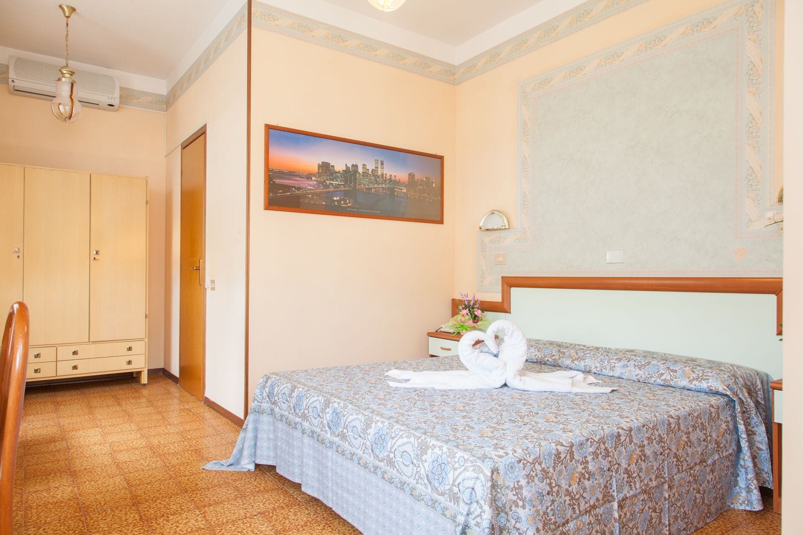 Una habitación de hotel con cama, armario, escritorio con lámpara y un cuadro de un paisaje urbano.