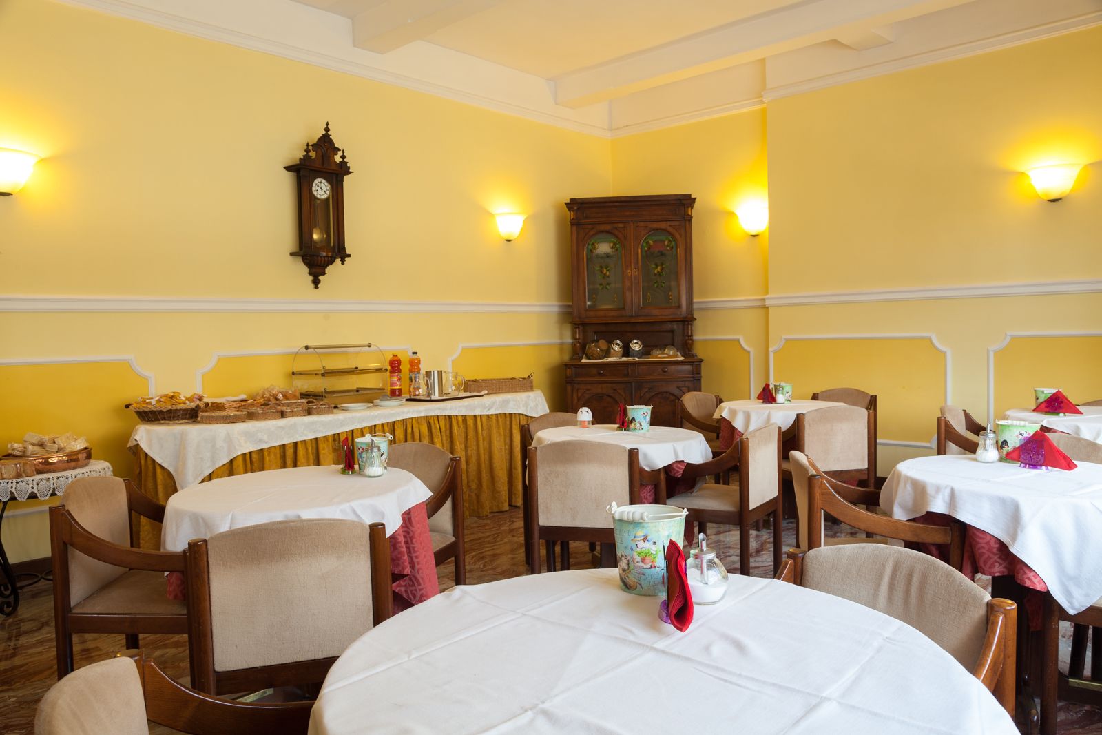 Un comedor con mesas redondas cubiertas con manteles blancos, sillas de madera y una mesa de buffet con alimentos.