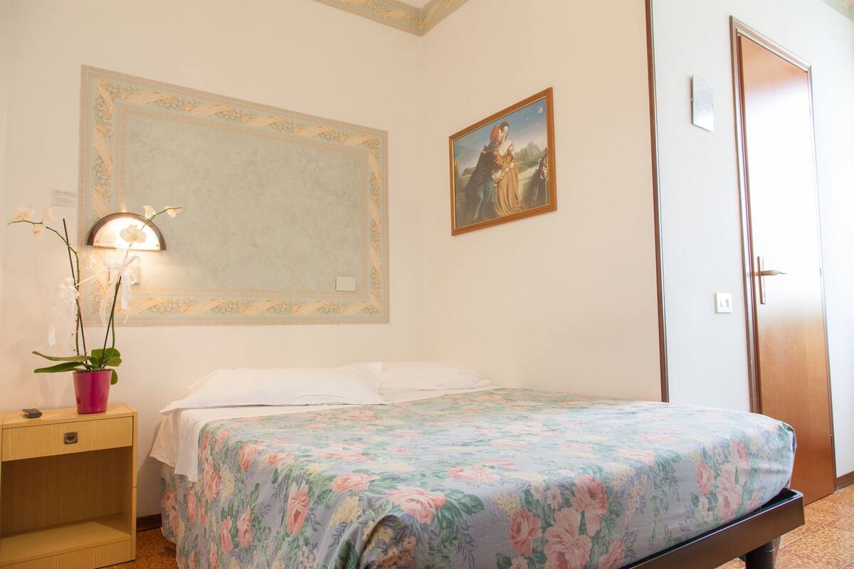 Une chambre d’hôtel avec un lit, une table de chevet, une plante en pot, un tableau et une porte.