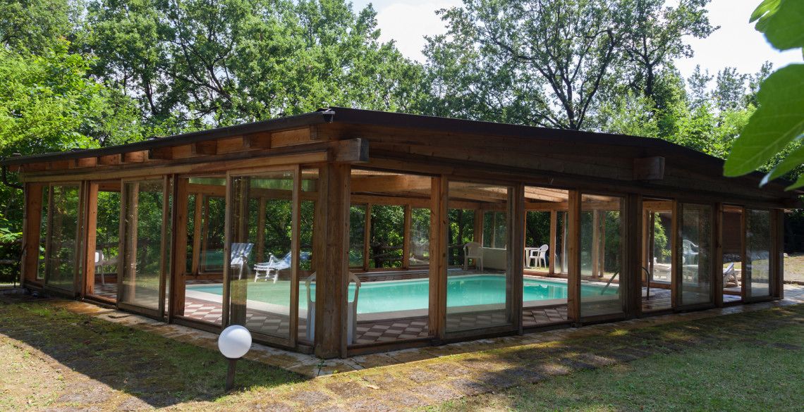 Une piscine extérieure couverte, dotée de parois en verre, entourée d’une terrasse en bois et d’une pelouse.