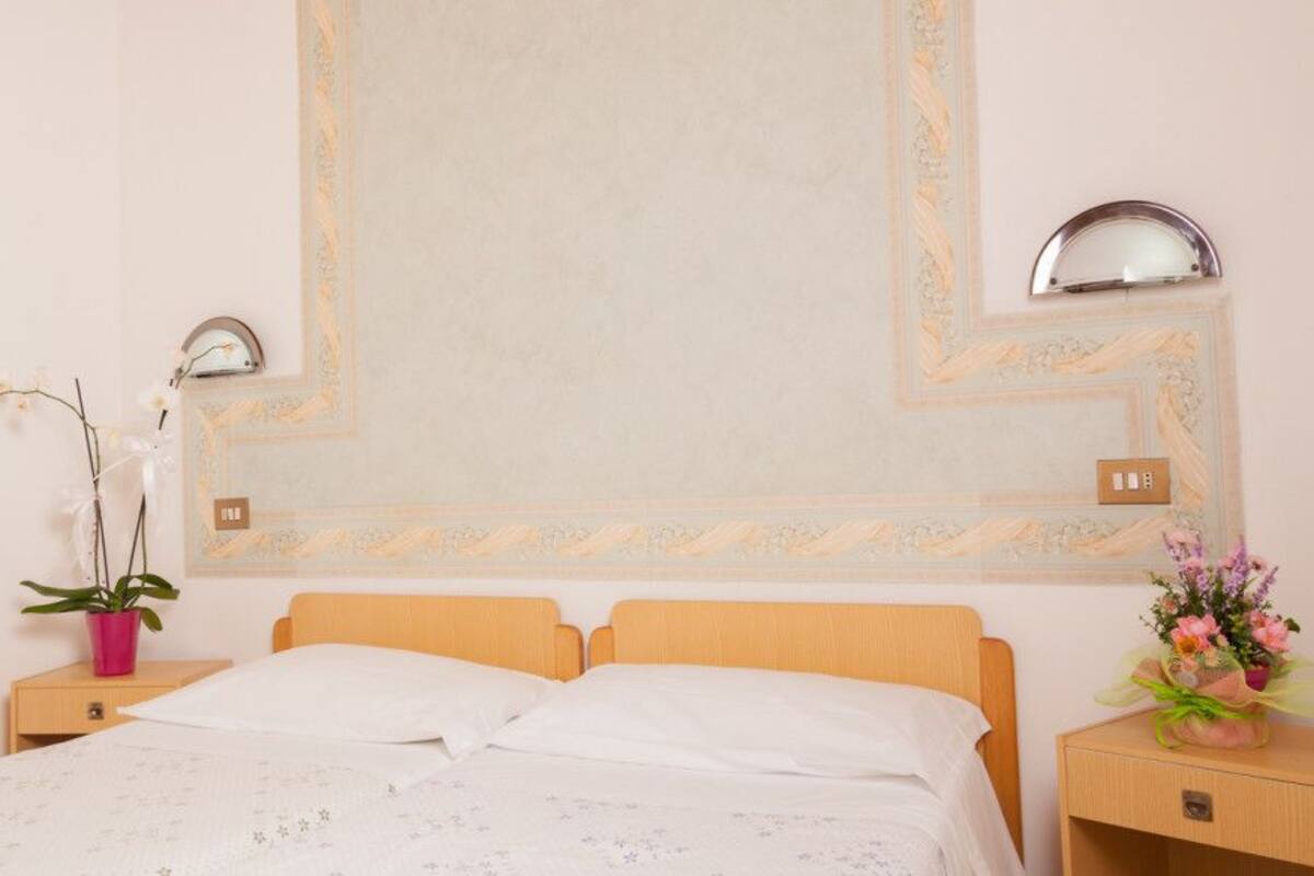 Un lit bien fait, avec une tête de lit en bois, une table de chevet ornée d’un vase avec une fleur rose, et un miroir fixé au mur.