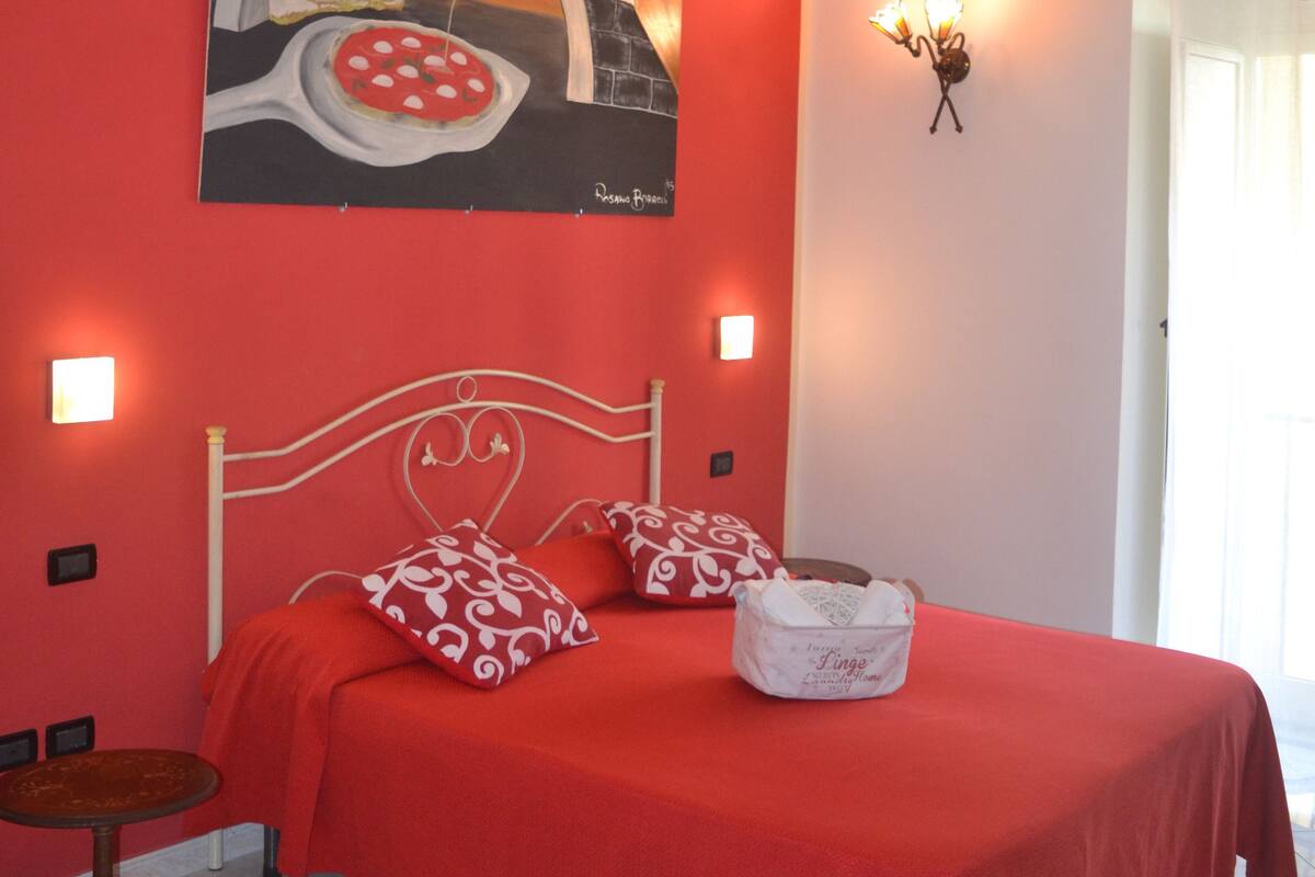 A bedroom with a red bedspread, a painting of a pizza oven, and a bedside table with a lamp.