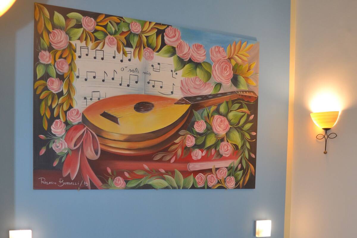 A hotel room with a bed, a painting of a guitar and roses, and two wall-mounted lamps.